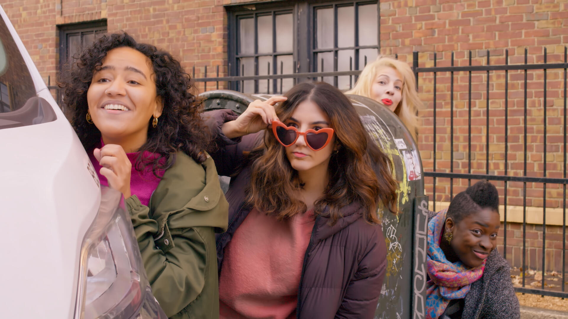 The four leading women of Late Bloomers lurk behind a car, watching intently across the street. They all look excited.