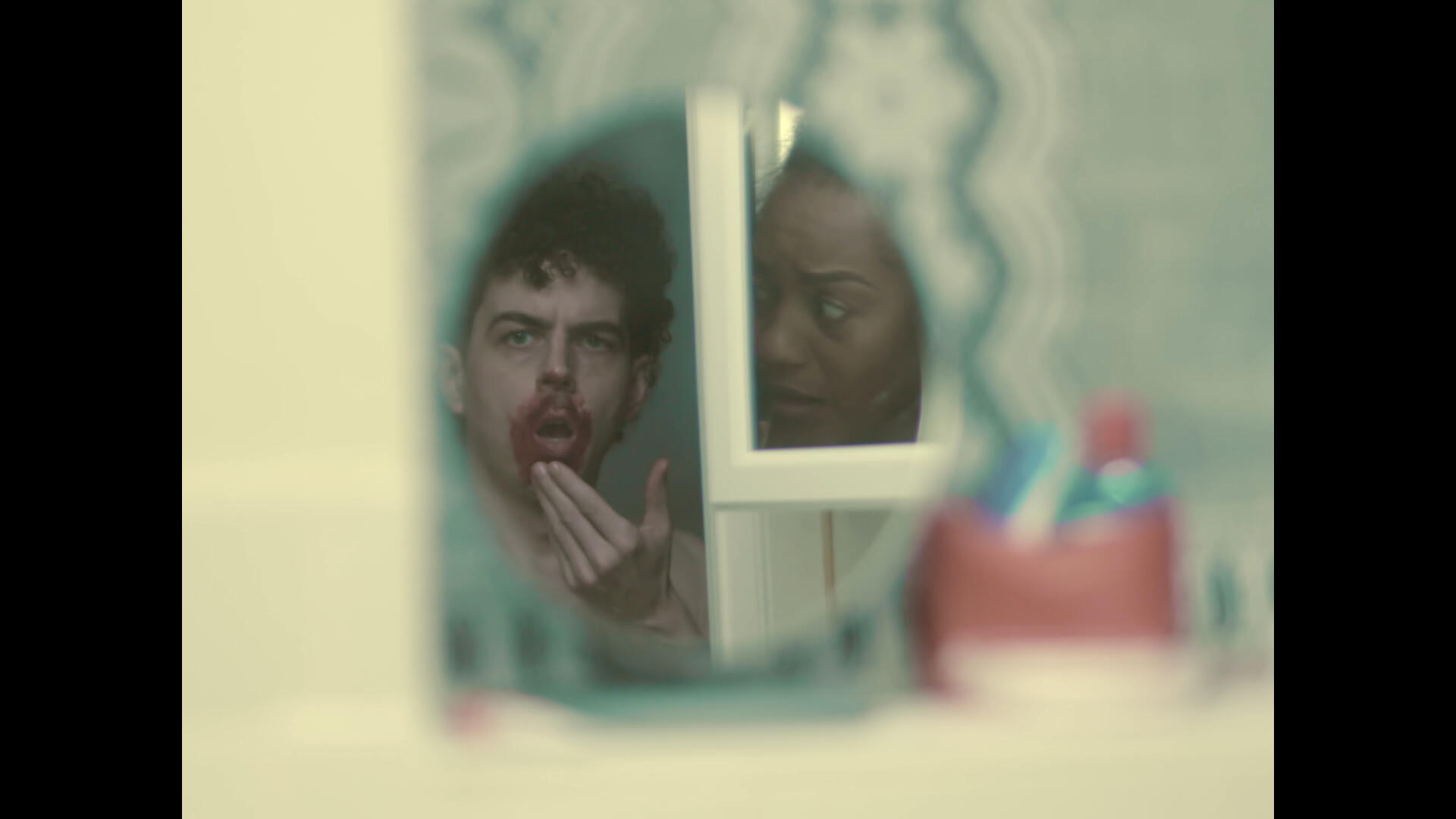 Period. Still 2 The leading couple stand in the bathroom. The lead actor looks in a mirror on the wall, observing blood around his mouth. The lead actress, reflected in the mirror on the medicine cabinet, looks mortified at her partner.