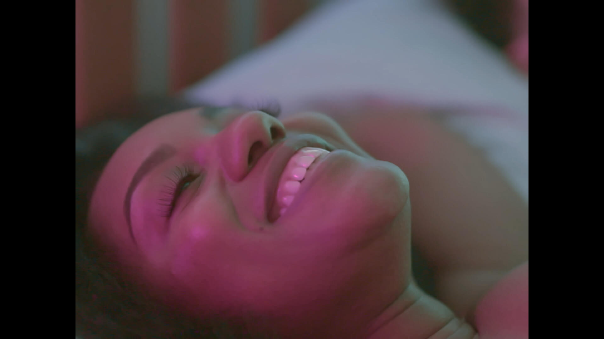 Period. Still 1 The leading actress lays back on a bed, smiling. She's bathed in green and magenta light.