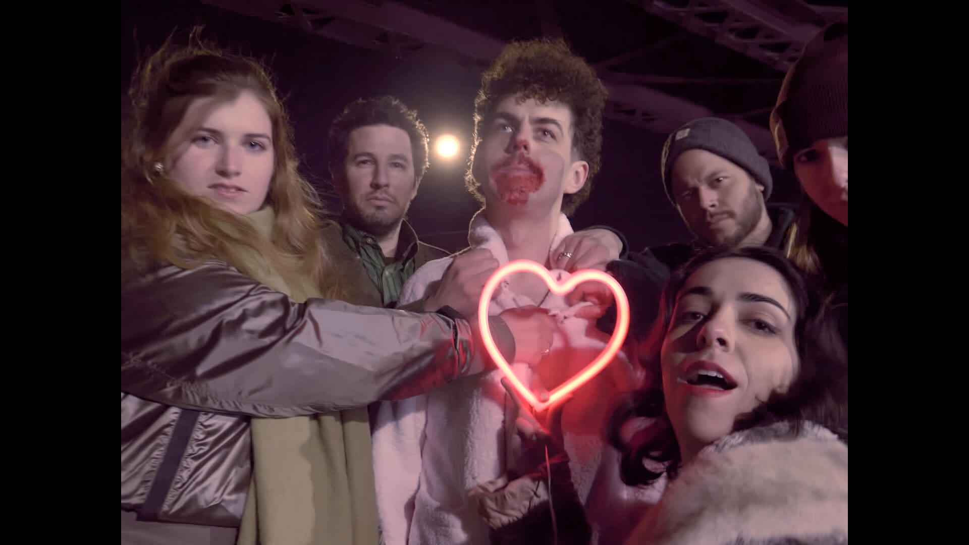 Period. Still 4 The lead actor stands on the street, blood on his mouth, surrounded by a group of people. One of them holds a neon-lit heart up to him.