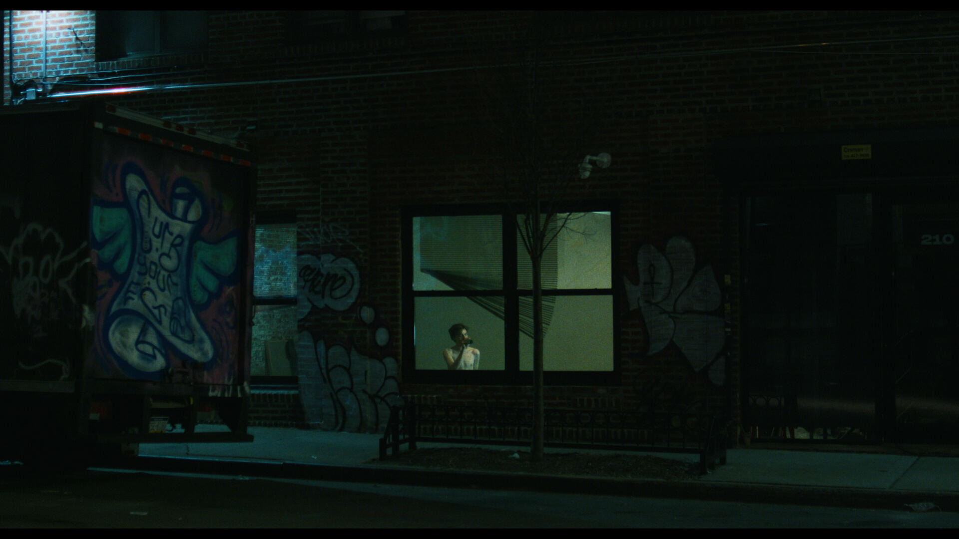 The antagonist of Working for the Weekend sits in a dimly lit room, as seen from across a dark street.